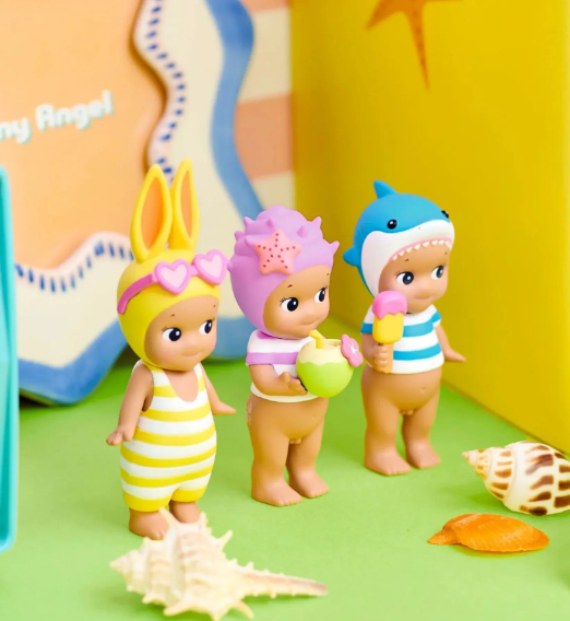 Three small toy baby figures dressed in pastel beach-themed outfits, standing on a green surface with seashells and a bright yellow backdrop.